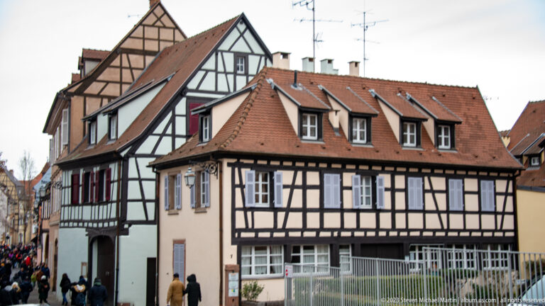 Black-and-white half-timbered houses with steep red tile roofs, dormers, and pedestrians at street level.