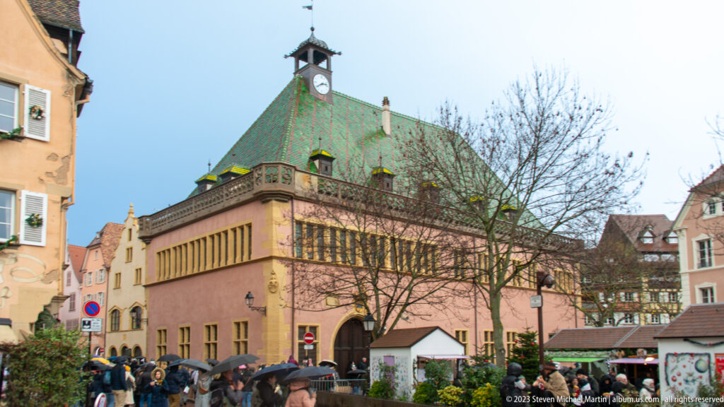 Koïfhus Old Customs House with a broad green-patterned roof, clock turret, masonry walls, and street activity below.
