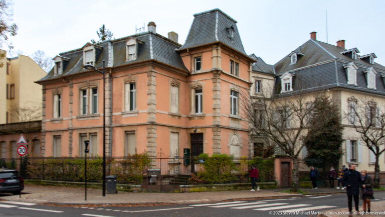 Salmon-colored historic villa with stone trim, tall windows, dormers, mansard roof, fence, and small front garden.