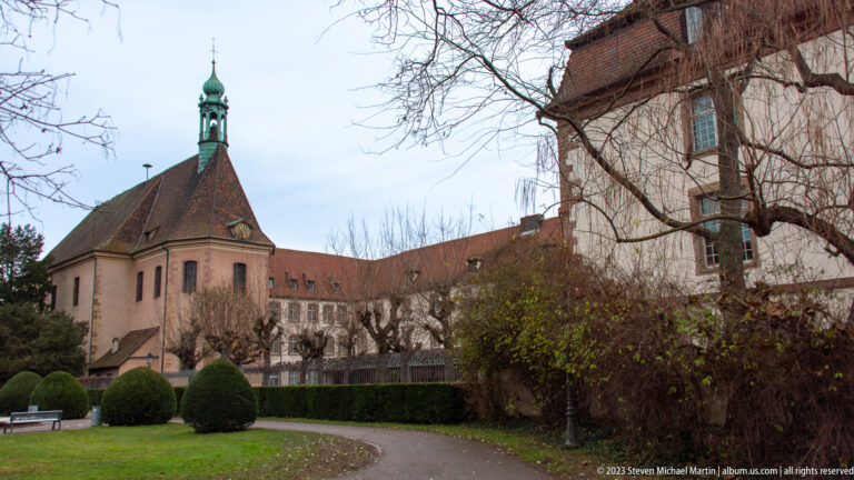 Historic church or convent buildings with tiled roofs and a small bell turret beside clipped garden shrubs and paths.
