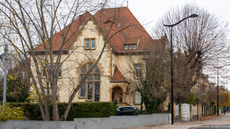 Cream historic villa with steep red-tiled roof, projecting gables, tall windows, and arched entrance behind winter trees.