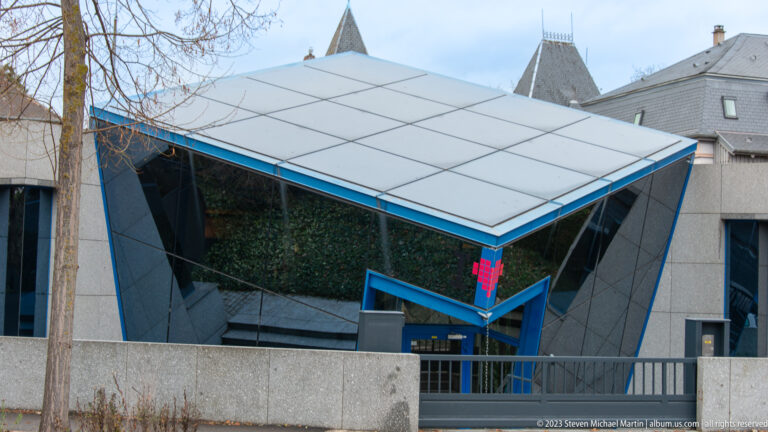 Angular modern entrance with reflective glass, white panels, blue framing, and red geometric emblem.