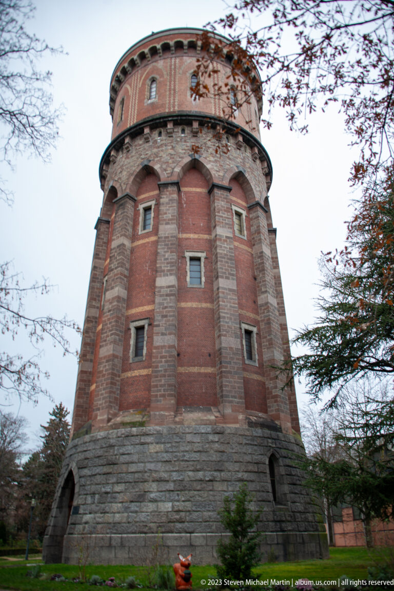 Round red-brick water tower with stone base, vertical masonry bands, small windows, and projecting circular cornice.