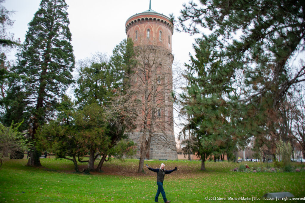 Tall cylindrical brick water tower rising above trees and lawn, with a person standing in the foreground for scale.