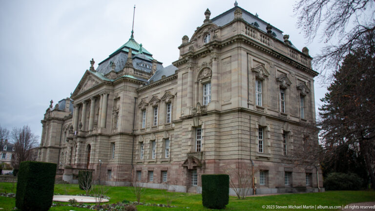 Large ornate stone civic building with pilasters, sculpted windows, balustrades, copper roof, lawn, and clipped hedges.