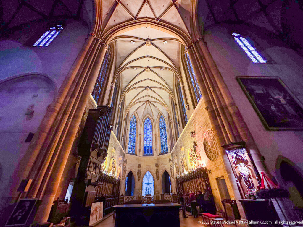 Gothic church nave with rib vaults, clustered columns, stained-glass choir windows, and dark side furnishings.