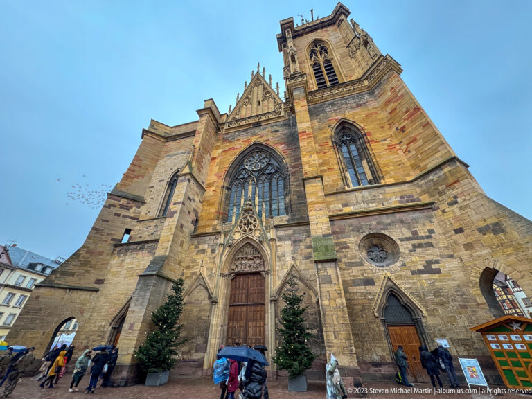Sandstone Gothic church façade with large traceried window, sculpted portal, unequal towers, and Christmas trees at the entrance.