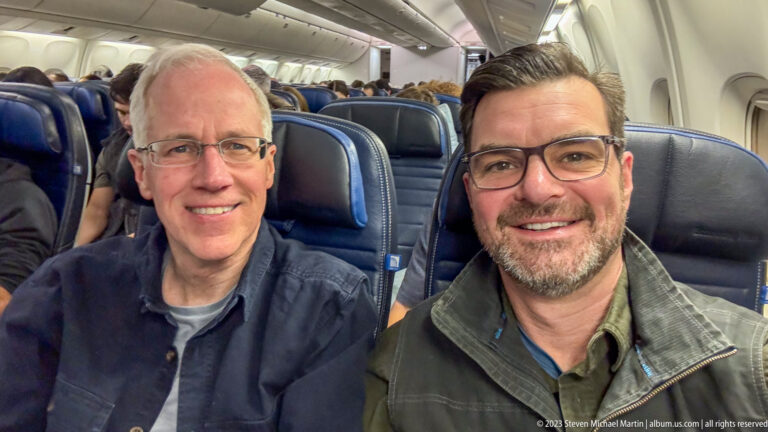 Two smiling men seated beside each other in blue airline seats inside an aircraft cabin.