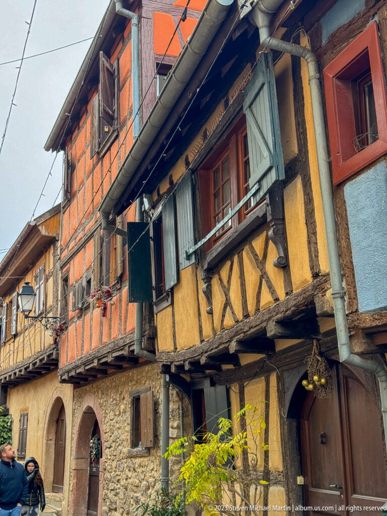 Ochre and orange half-timbered houses with open shutters and projecting upper floors along a narrow lane.