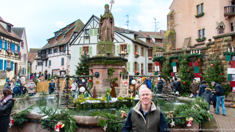 Smiling visitor standing before a Christmas-decorated fountain topped by a stone saint statue in a crowded Eguisheim square.