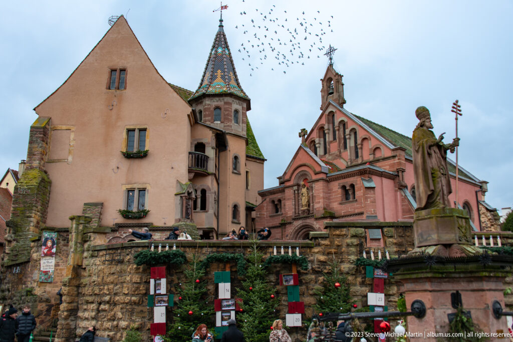 Stone saint statue beside Christmas decorations with a pink church, patterned-roof turret, historic walls, and birds overhead.