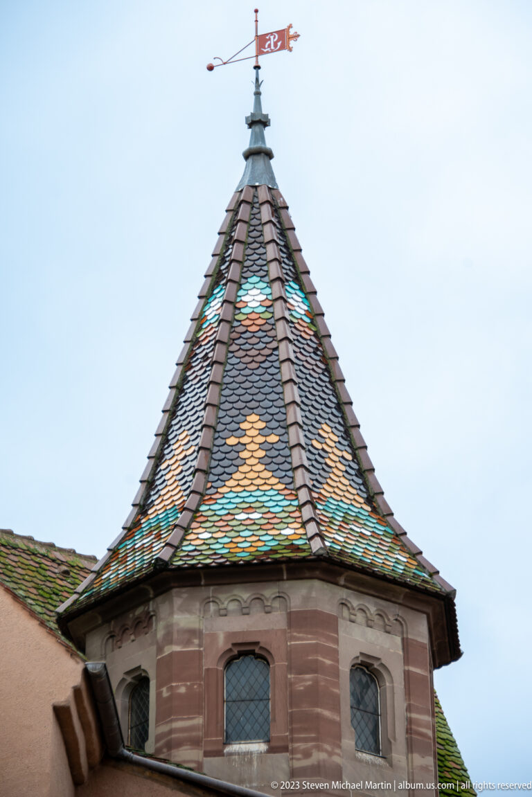 Polygonal turret with multicolored patterned tile roof, pink stone walls, leaded windows, finial, and weather vane.