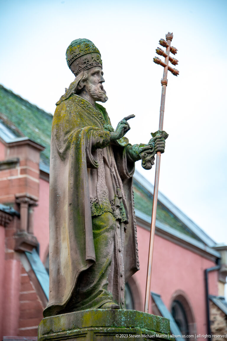 Weathered stone statue of a robed ecclesiastical figure holding a tall staff, with pink church architecture behind.