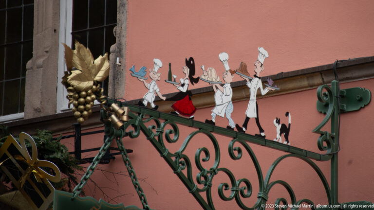 Painted cutout cooks and figures marching above ornate green wrought iron with a gilded grape cluster.