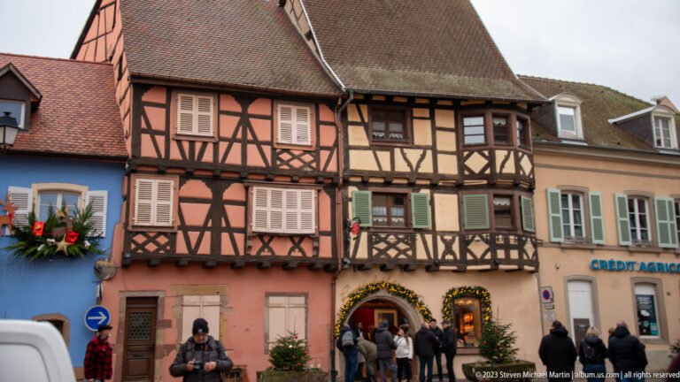 Pink and brown half-timbered façades beside a Crédit Agricole building, with pedestrians and Christmas decorations.