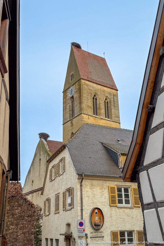 Square stone church tower with paired arched openings rising above houses in central Eguisheim.
