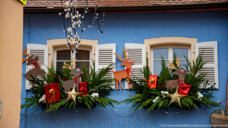 Two white-shuttered windows on a blue façade with evergreen boxes, wooden reindeer, red gifts, stars, and lights.