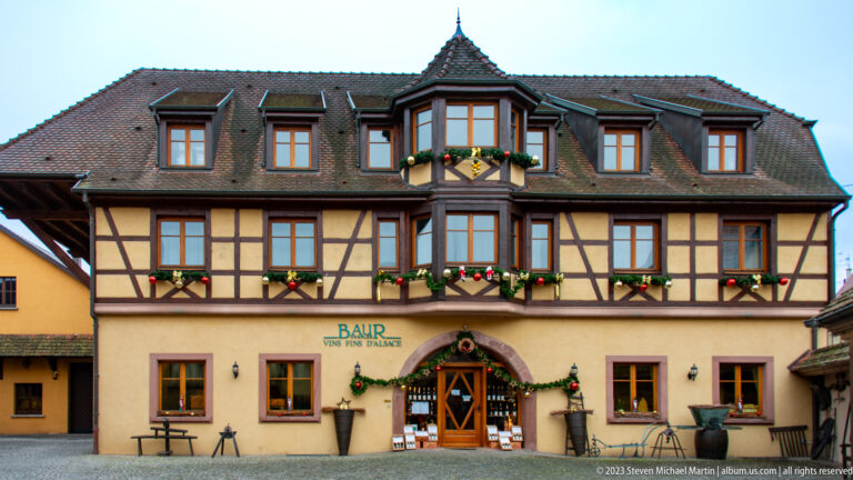 Baur Vins fins d’Alsace half-timbered building with central bay, dormers, flower boxes, and decorated arched entrance.
