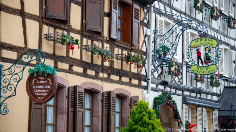 Ornate hanging signs for Hebinger Vins d’Alsace and Auberge Alsacienne projecting from decorated half-timbered façades.
