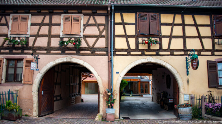 Two rounded stone passage arches beneath adjoining half-timbered houses, one with heavy wooden doors partly open.