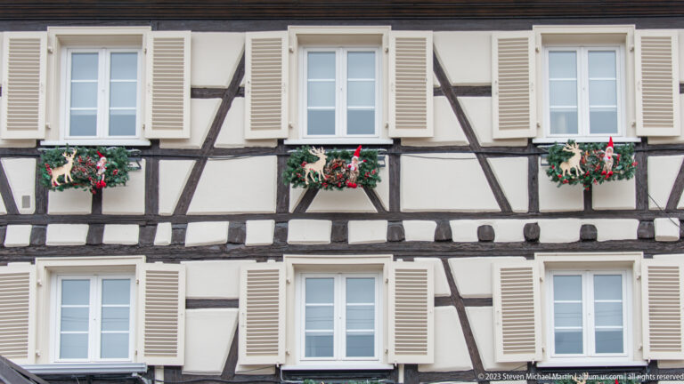 Half-timbered pale façade with six white-shuttered windows and Christmas greenery beneath several openings.