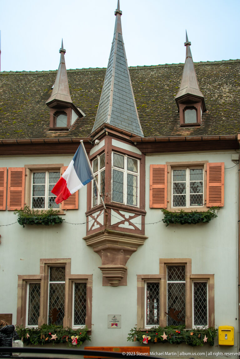 Pale historic building with French flag, projecting polygonal turret, orange shutters, diamond-paned windows, and tiled roof.