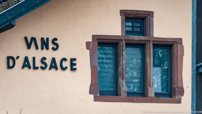 “Vins d’Alsace” lettering beside three narrow reddish-stone-framed windows with blue-green coverings.