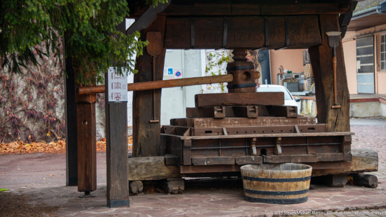 Large traditional wooden wine press with screw mechanism beneath a timber shelter, with a small wooden tub in front.