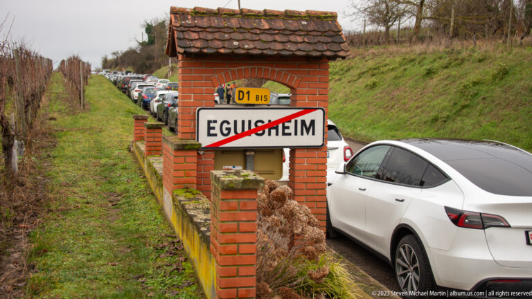 End-of-town Eguisheim road sign on D1 bis beside a line of cars and grassy roadside embankments.