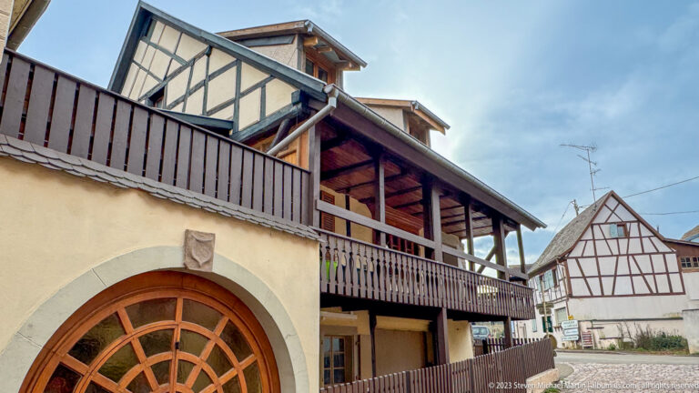 Pale historic house with a large arched wooden door and long dark wooden gallery above.