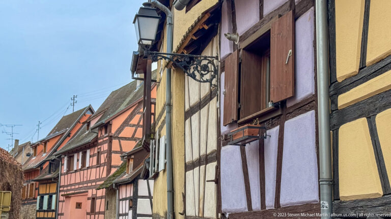 Oblique row of colorful half-timbered houses with shutters and a wrought-iron lamp bracket along a narrow street.