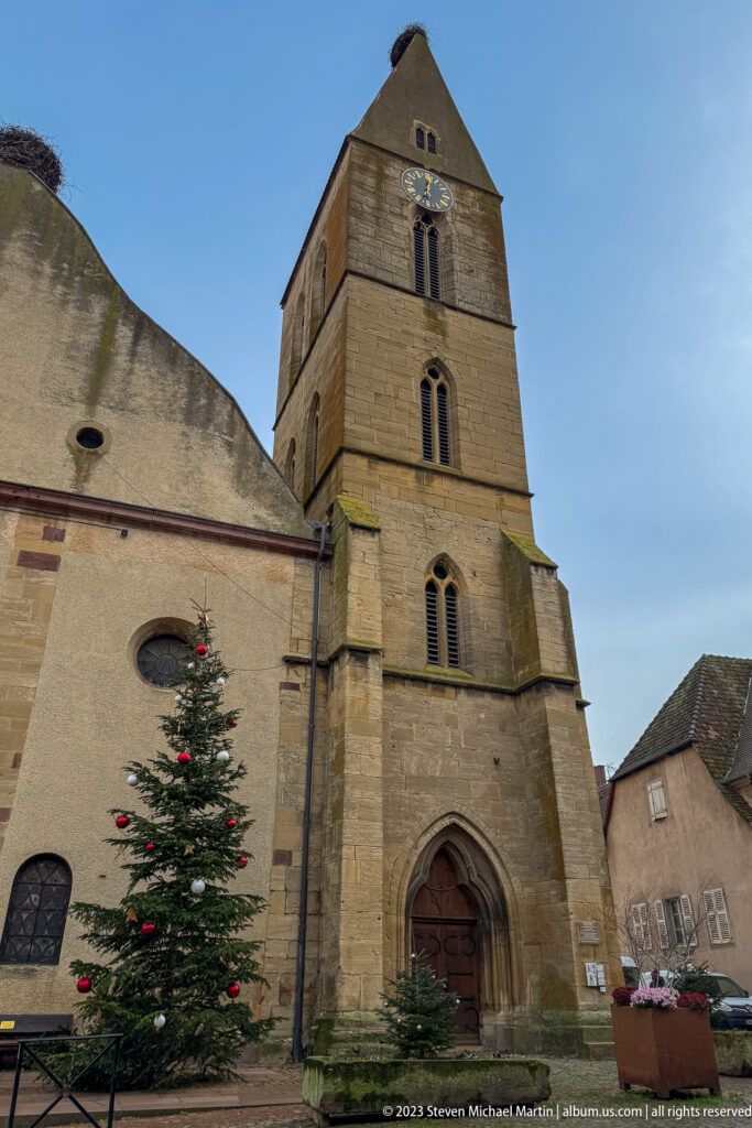 Stone parish church with a massive square tower, pointed doorway, buttresses, and a decorated Christmas tree outside.