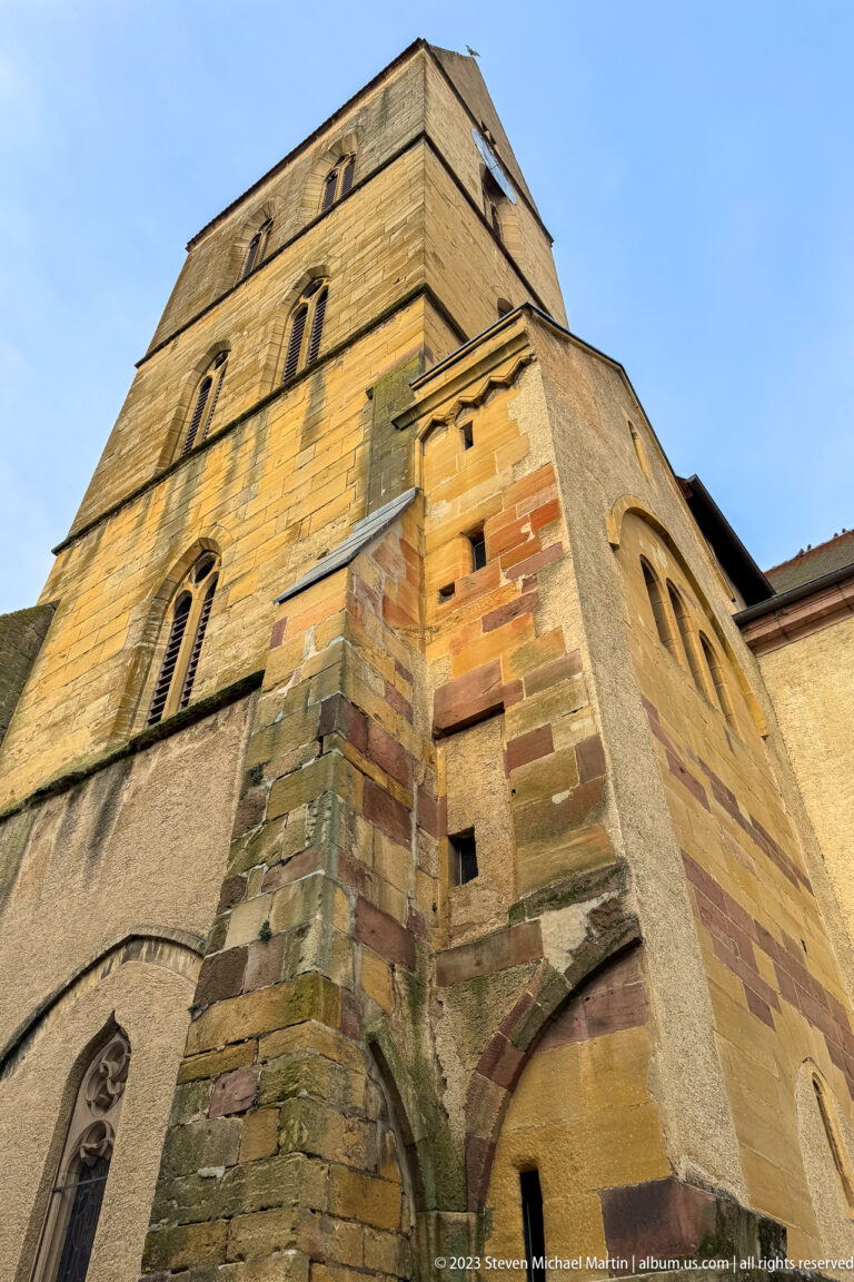 Low-angle view of a stone church tower, projecting buttress, and narrow arched openings against blue sky.