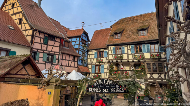 Christmas-decorated courtyard among half-timbered houses, with a rustic Vin Chaud sign and seasonal figures.