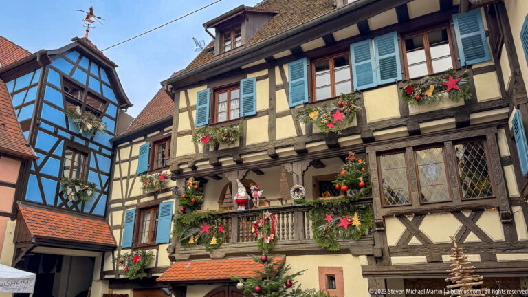 Half-timbered house with blue shutters, balcony, leaded windows, garlands, red stars, and Christmas decorations.