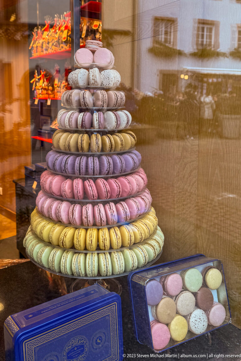 Tiered pyramid of pastel macarons displayed behind a reflective shop window with boxes below.