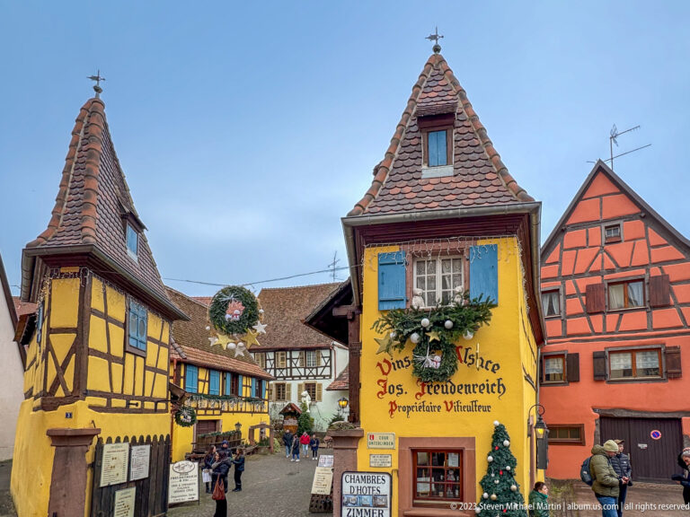 Yellow wine merchant building with blue shutters, Christmas greenery, and painted Vins d’Alsace Jos. Freudenreich lettering.