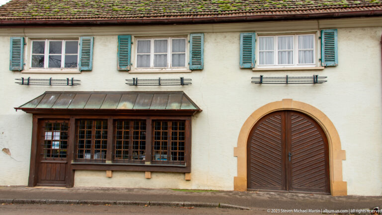 White historic façade with green shutters, a dark glazed opening, and a large round-arched wooden carriage door.