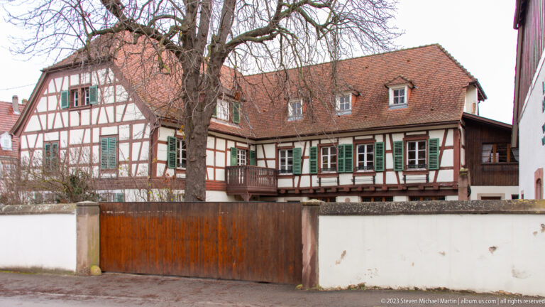 Large half-timbered house with green shutters behind a white wall, wooden gate, and leafless courtyard tree.