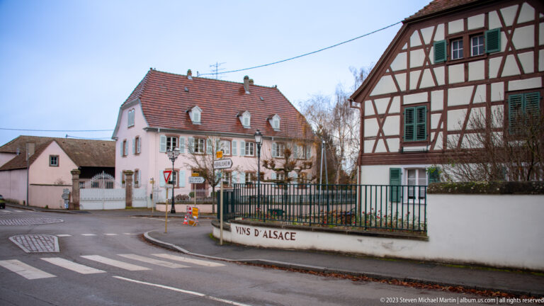 Road junction beside a fenced wine property, with a pink house and large half-timbered residence behind a Vins d’Alsace wall.