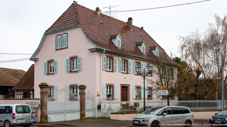 Large pink corner house with pale shutters, dormers, red tiled roof, metal fence, and parked cars.
