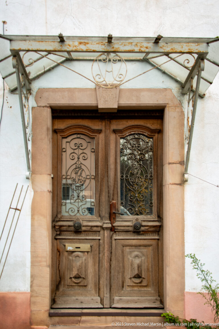 Weathered double wooden door with glazed upper panels, wrought-iron scrollwork, stone surround, and metal canopy.