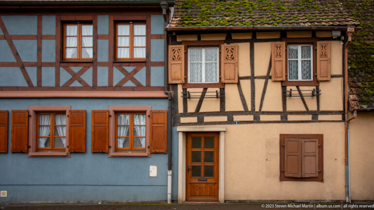 Blue and ochre half-timbered houses with orange and carved wooden shutters, closely framed at street level.