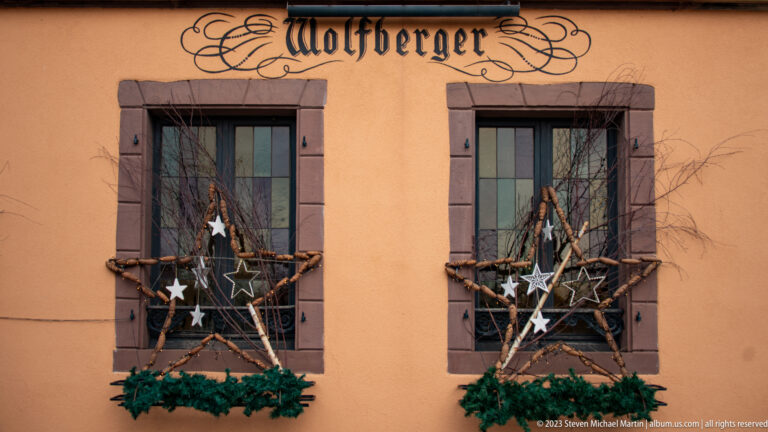 Two windows decorated with twig stars and evergreen swags beneath painted Wolfberger lettering on an orange wall.