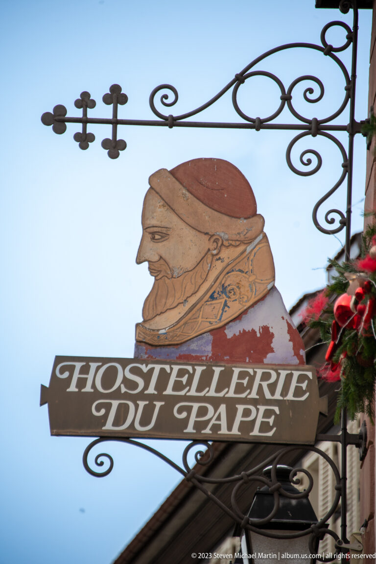 Wrought-iron Hostellerie du Pape hanging sign topped by a painted clerical profile against the sky.