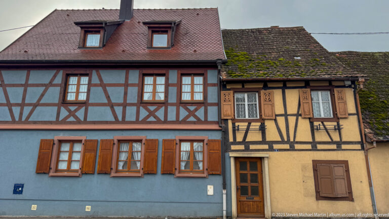 Blue and ochre half-timbered houses with shuttered windows and tiled roofs stand side by side.