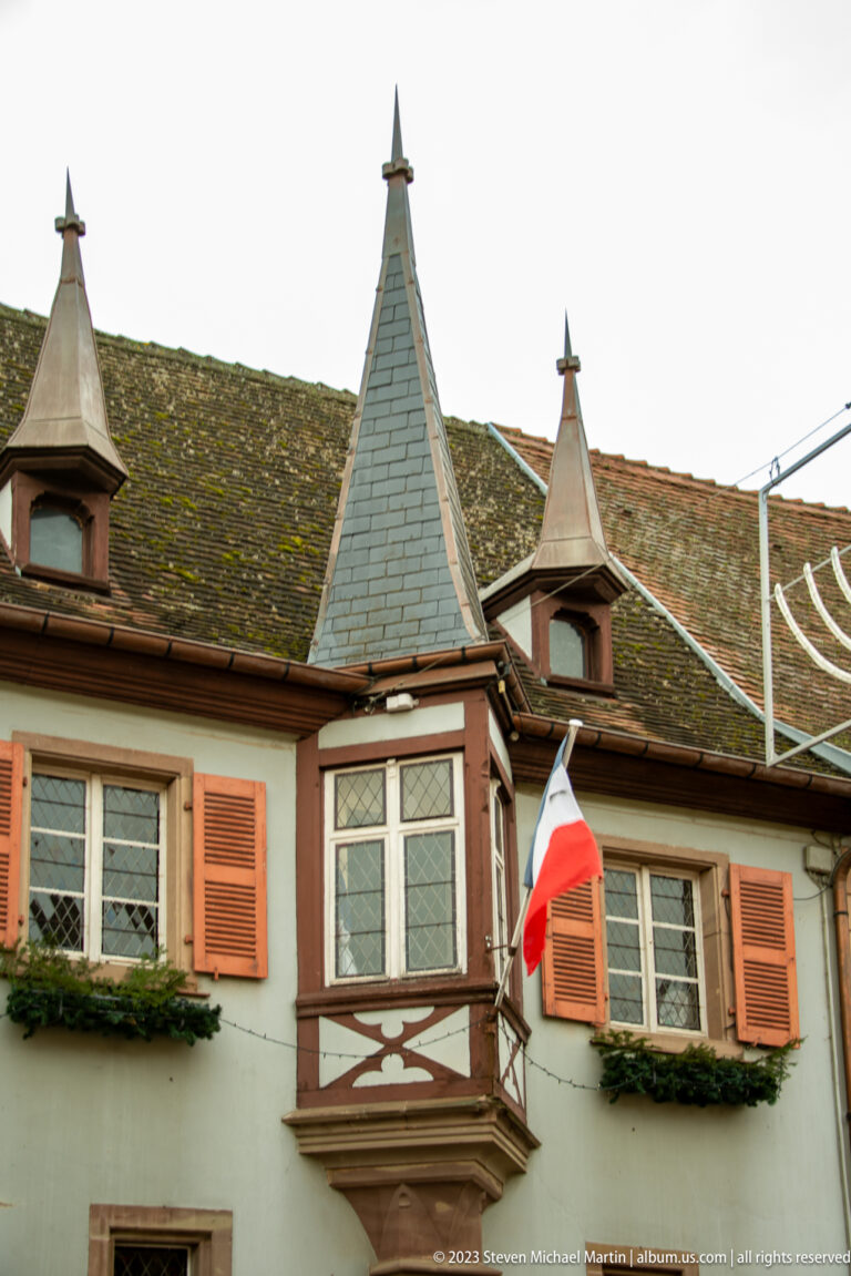 Historic house with orange shutters, pointed turret roofs, projecting bay, mossy tiles, and a French flag.