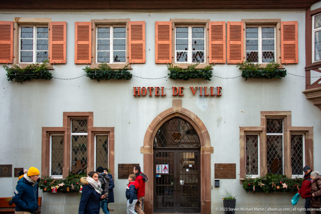 Eguisheim town hall façade with shuttered windows, flower boxes, arched doorway, and pedestrians gathered along the street.