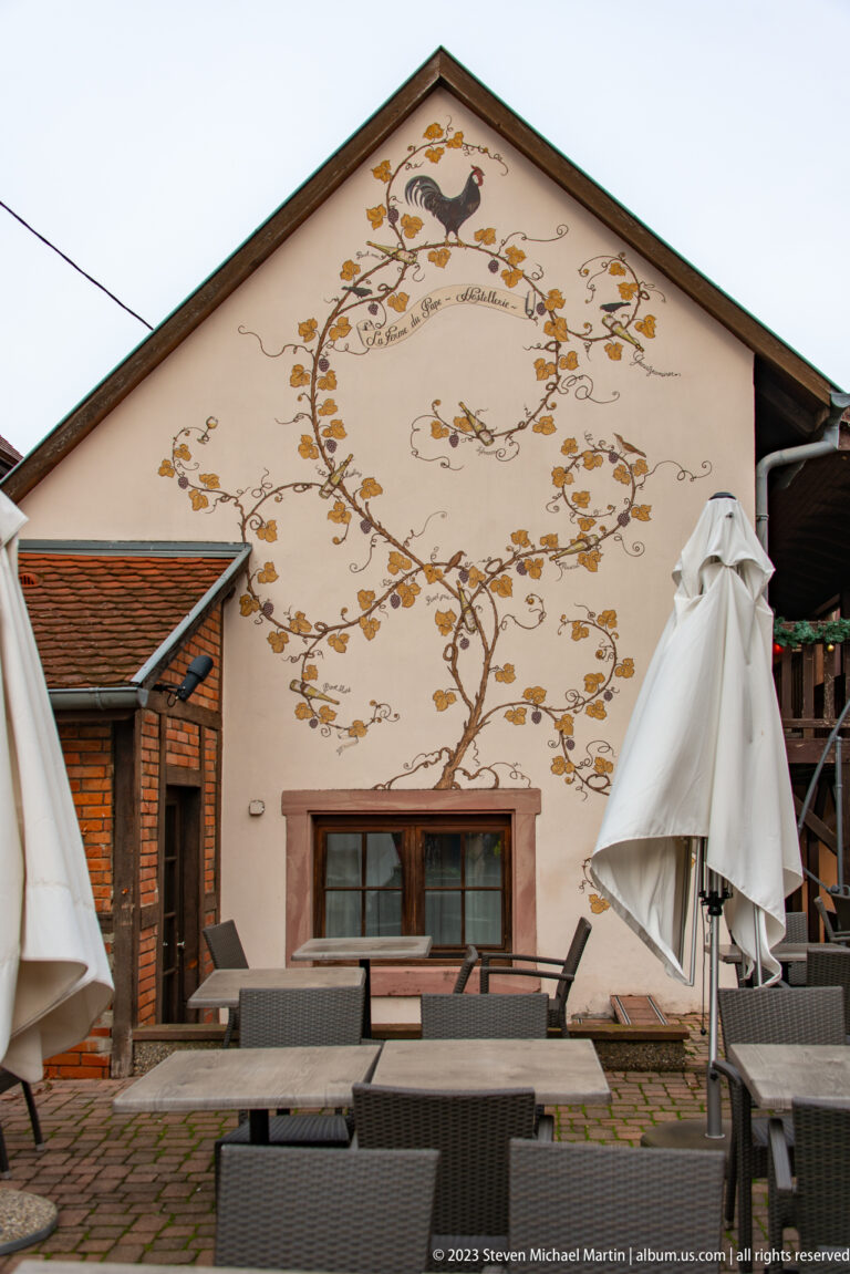 Painted gable wall with ochre scrollwork and floral motifs behind terrace furniture and folded umbrellas.