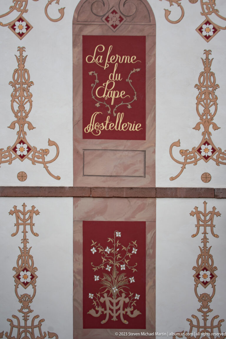 Decorated wall with “La Ferme du Pape – Hostellerie” sign, gold scrollwork, and red ornamental panels.